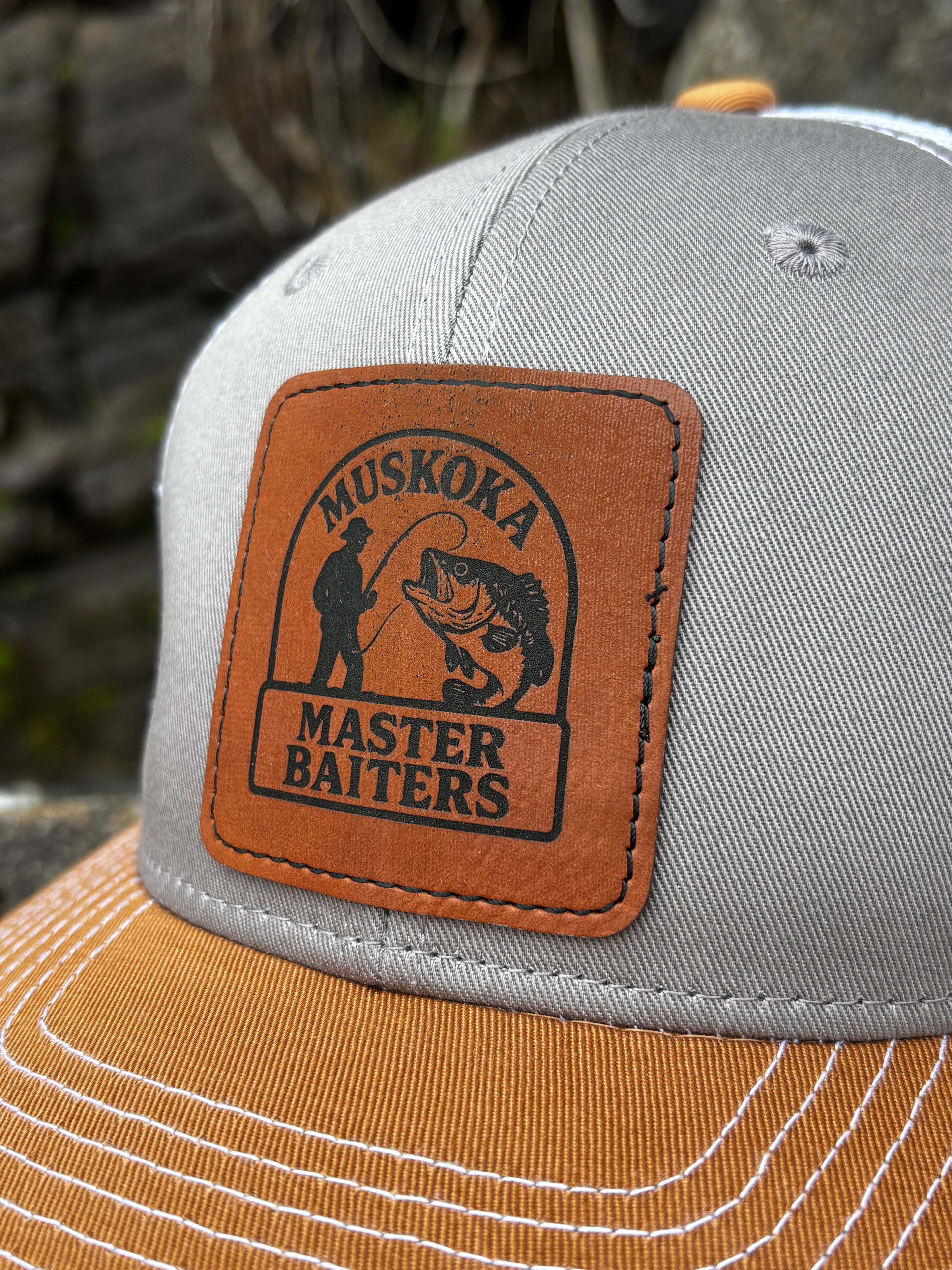Gray, duck brown and white mesh back trucker hat, with a Muskoka Master Baiter leatherette patch, on a blurred natural background.