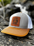 Gray, duck brown and white mesh back trucker hat, with a Muskoka Master Baiter leatherette patch, on a rocky surface.