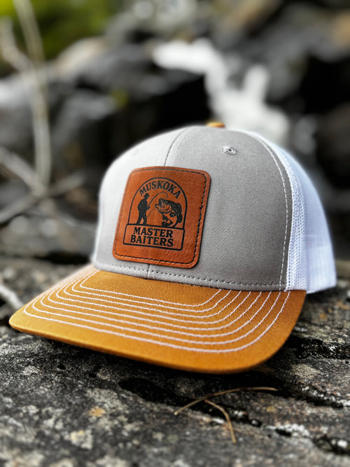 Gray, duck brown and white mesh back trucker hat, with a Muskoka Master Baiter leatherette patch, on a rocky surface.