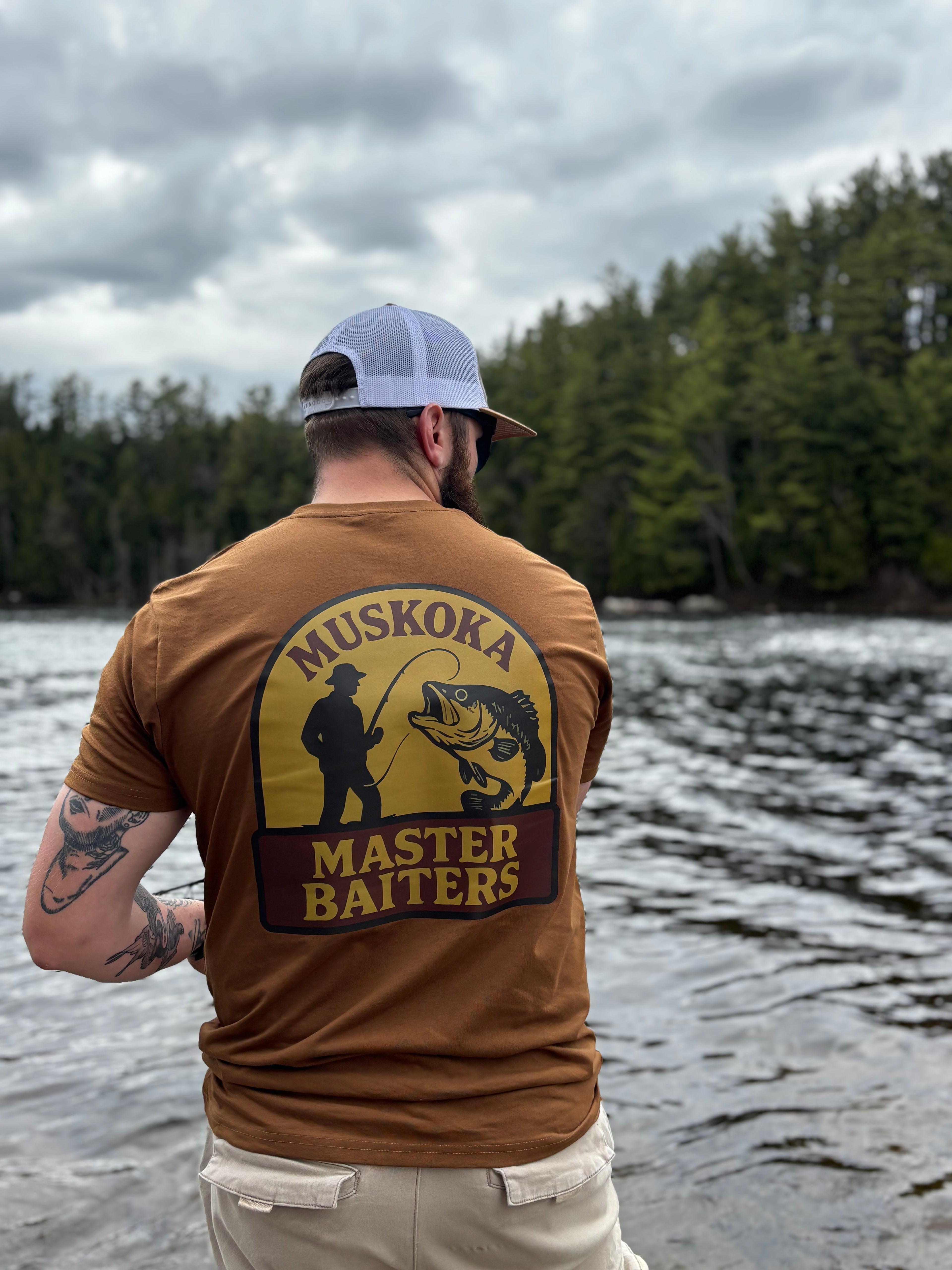 Man wearing a caramel 'Muskoka Master Baiters' t-shirt by a body of water with trees in the background.
